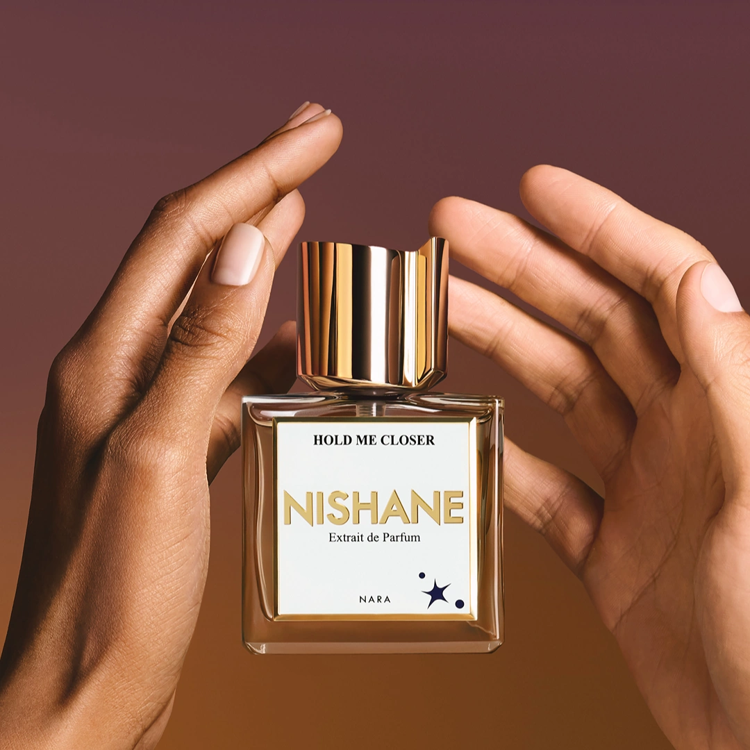 Nishane Hold Me Closer perfume bottle framed by two hands against a warm brown background.